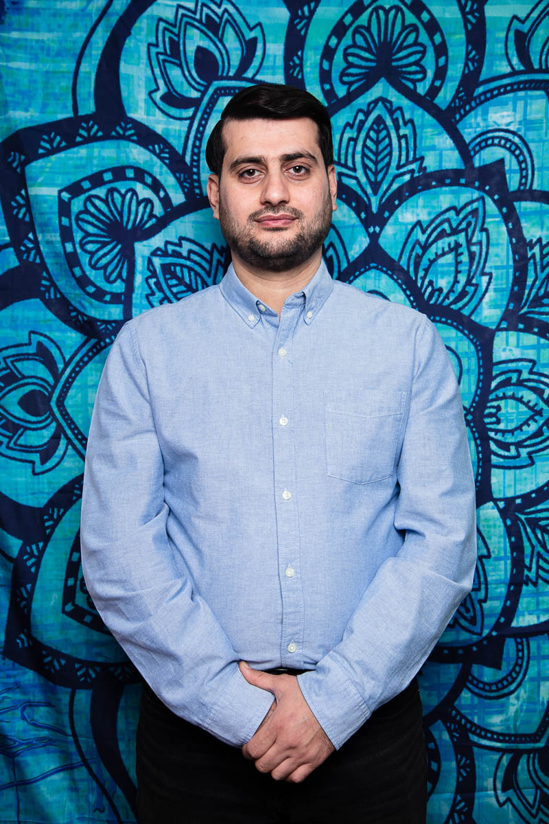 Portrait of refugee Sangar standing with his left hand clasping the right against a blue background with designs