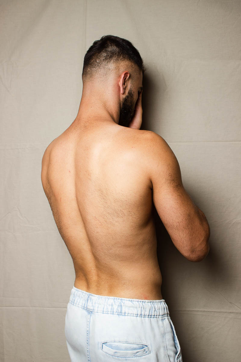 Portrait of refugee David shirtless with his back turned to the camera and his hinds hiding his face