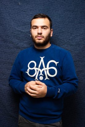 Portrait of refugee Mohamed standing against a denim blue background with his hands interlocked