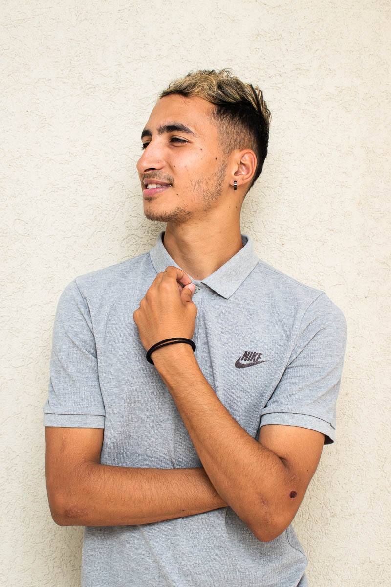 Portrait of refugee Lahcen looking to his right and smiling with a hand touching his collar and the other folder across his waist