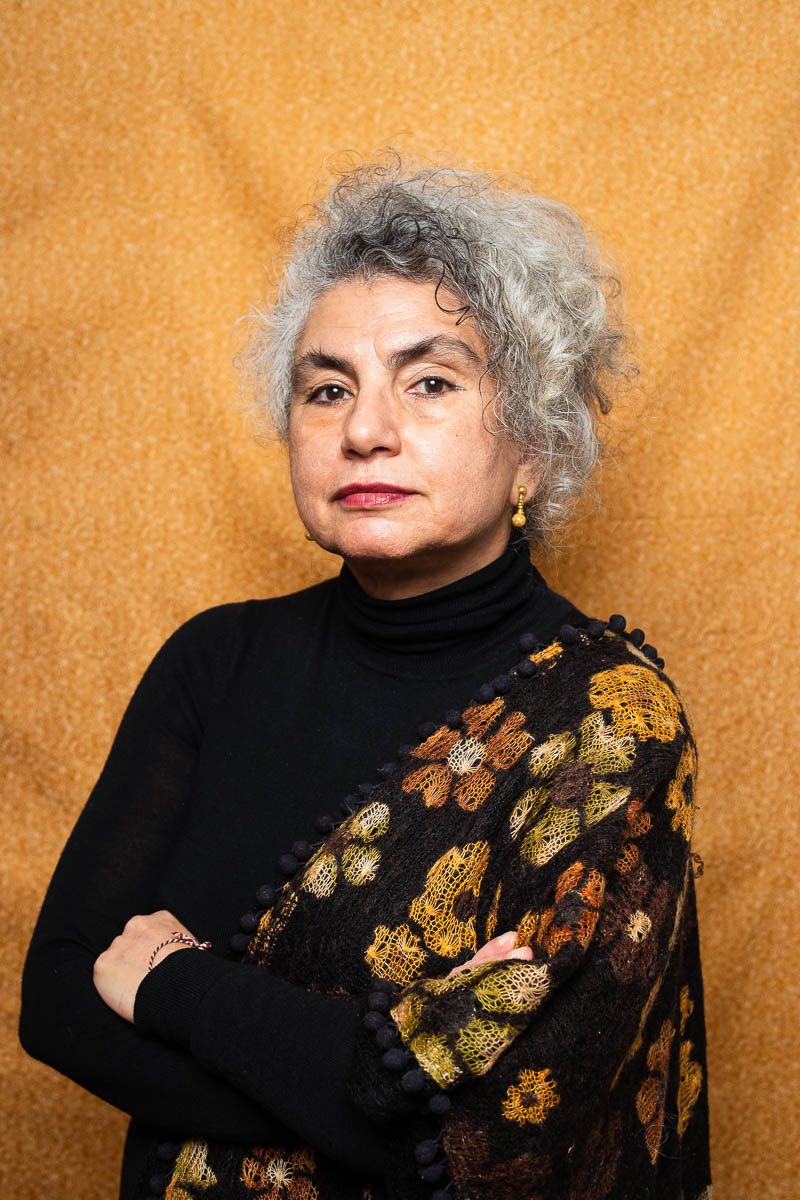 Portrait of refugee Banu with short graying hair and her hands folded across wearing a patterned shawl