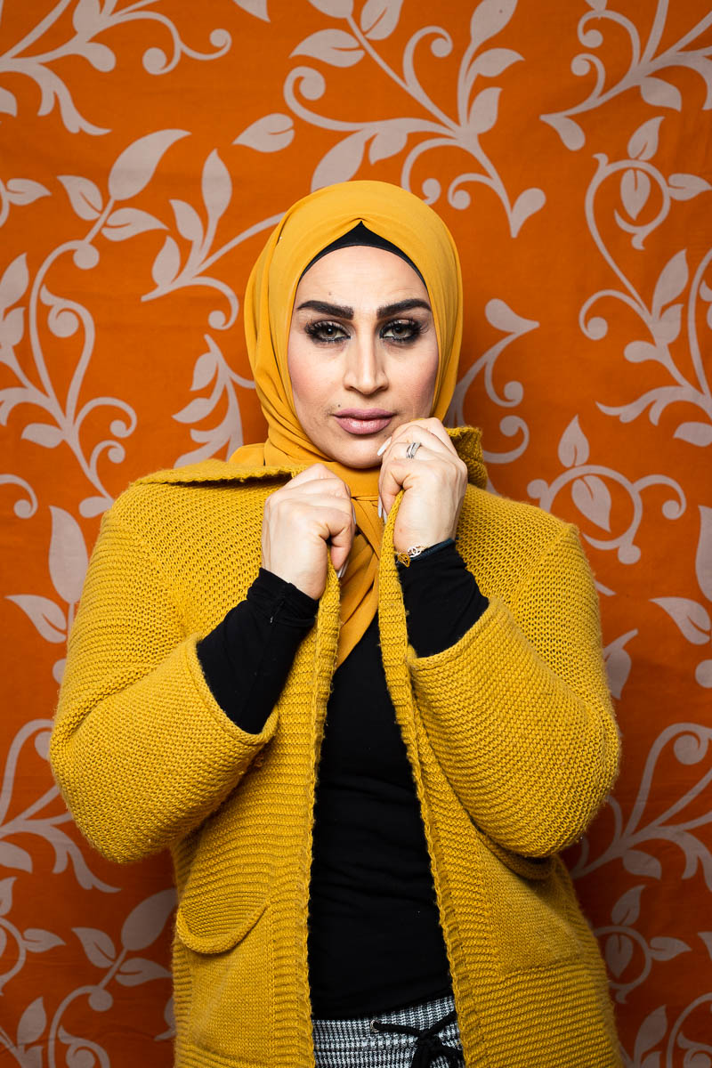 Portrait of refugee Rowaida wearing a mustard colored hijab and sweater while holding the collar of her sweater with both her hands