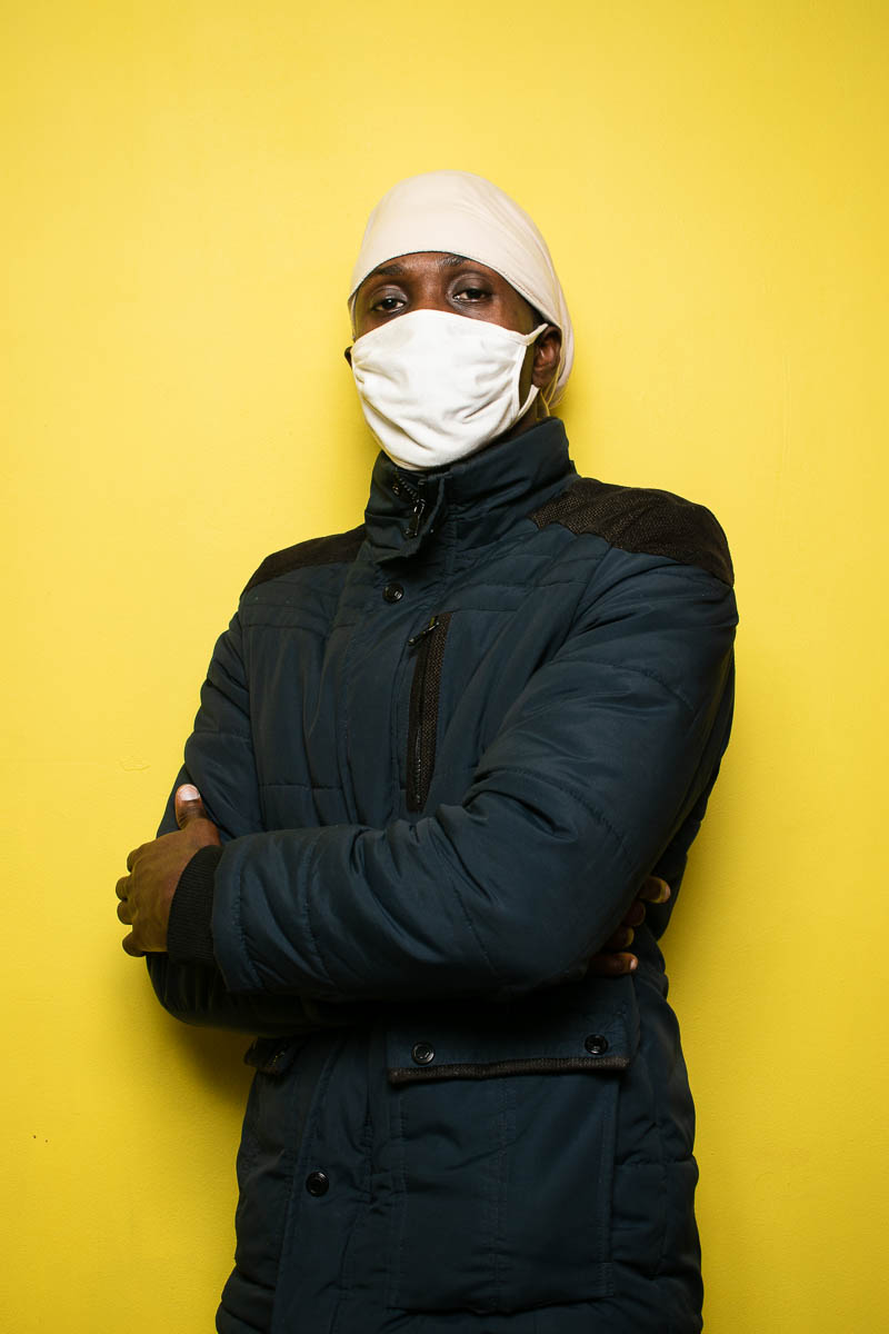 Portrait of refugee Martin wearing a jacket , white face mask and coving his heard with a white cloth standing with hands crossed against a yellow background