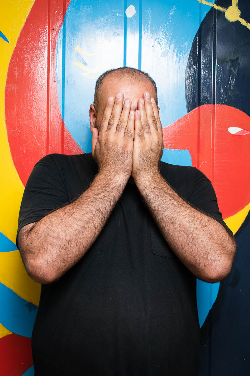 Portrait of refugee Parsa hiding his face with his hands standing against a colourful painted wall