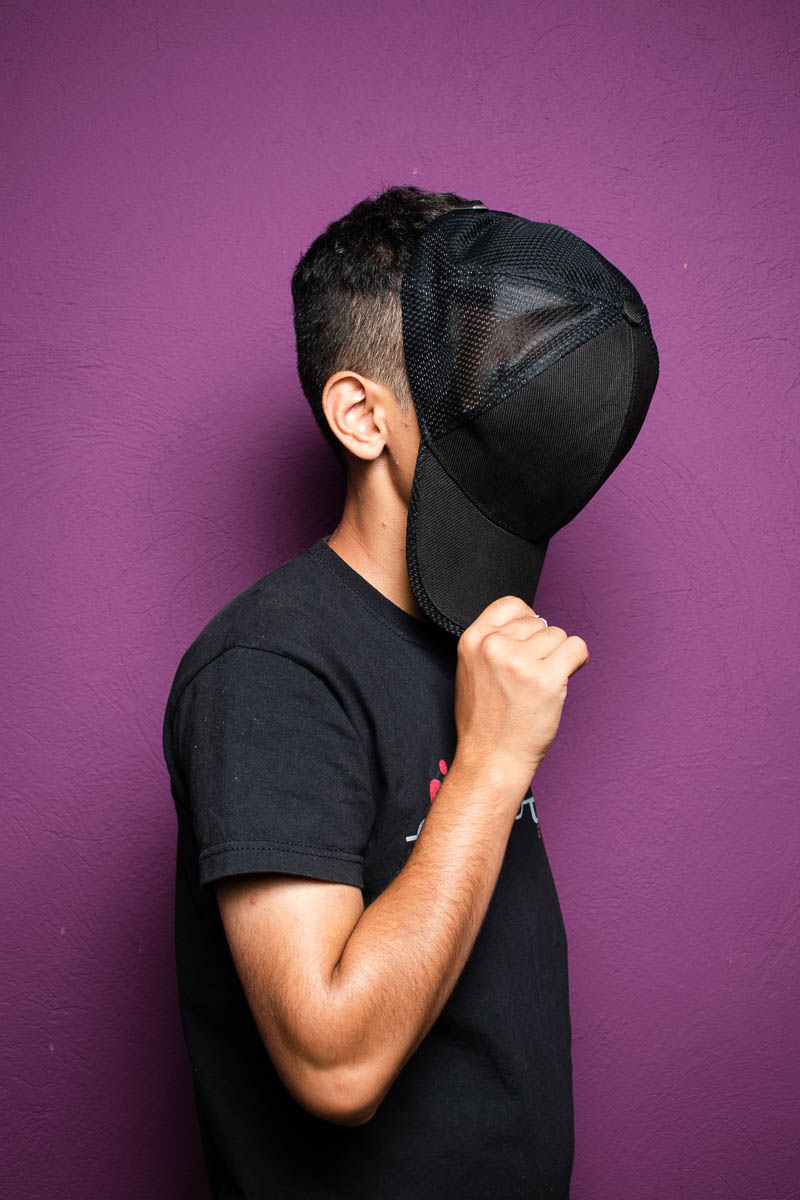 Portrait of refugee Ali standing sideways against a purple backdrop holding a snapback cap to cover his face