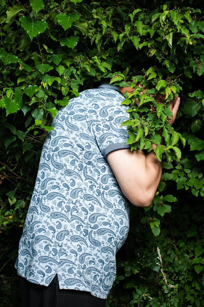 Portrait of refugee Ehsan wearing a blue printed shirt and hiding his face with the dense plant background