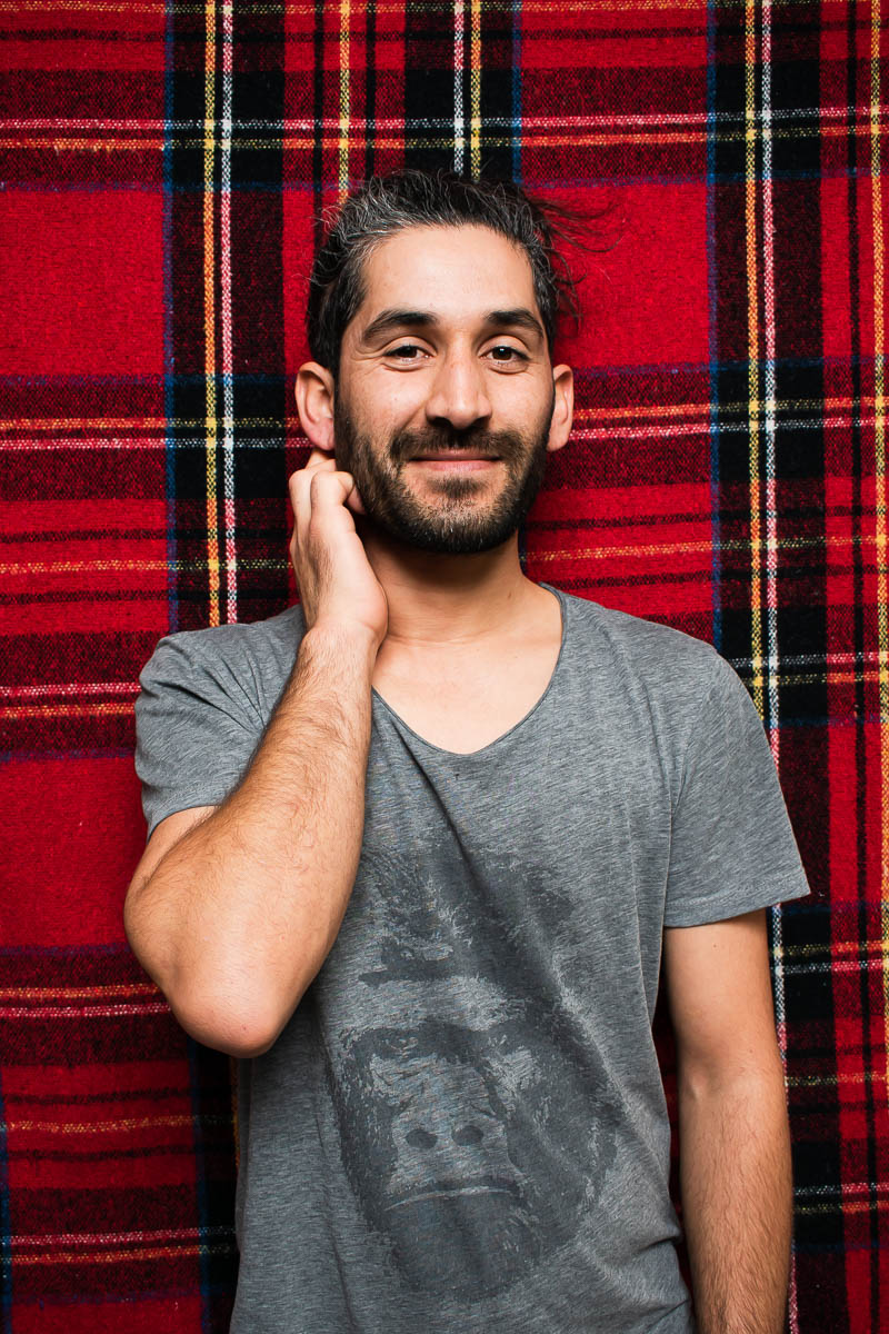 Portrait of refugee Hüseyin against a plaid background with his hand scratching his neck