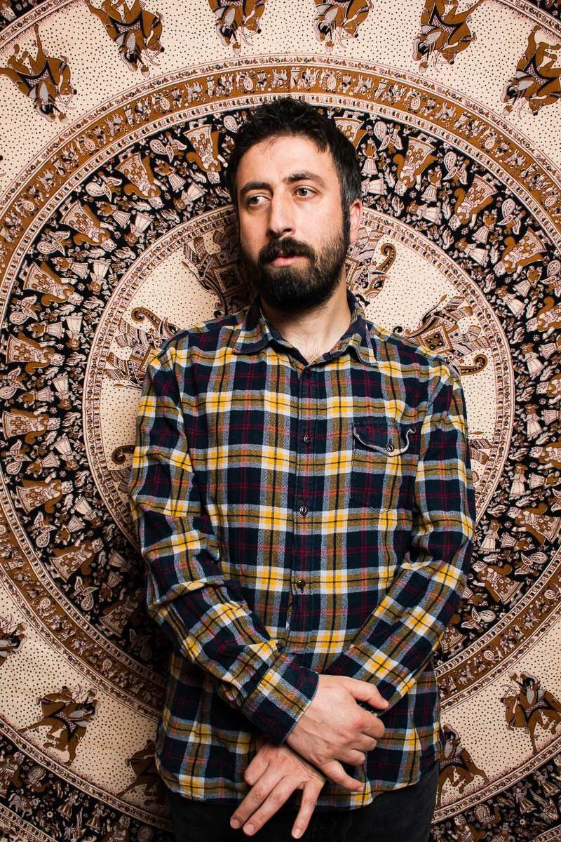 Portrait of refugee Mevlan wearing a plaid shirt standing with his hands clasped down against a printed mandala background