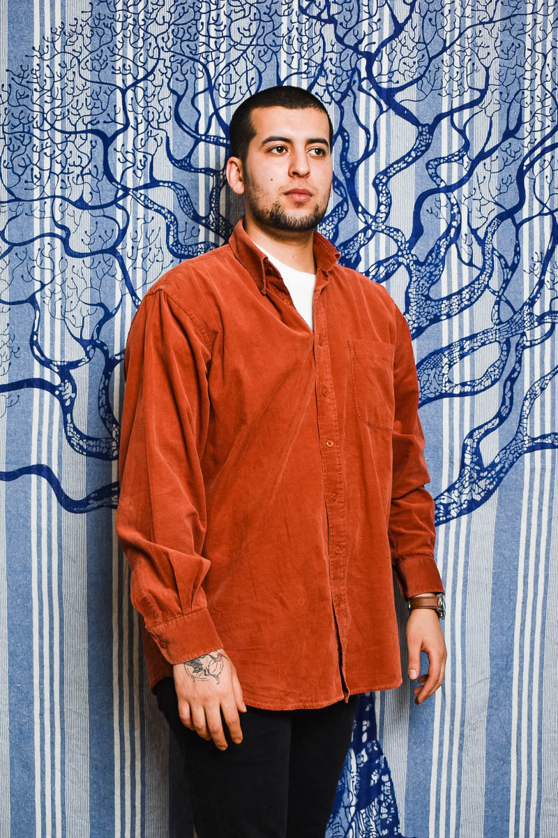 Portrait of refugee İsmail with a bird tattoo on top of his right hand standing against a background with a tree painted on it