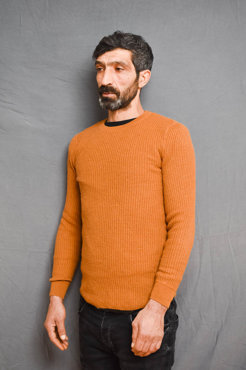 Portrait of refugee Zeki standing facing to his right in an orange sweater