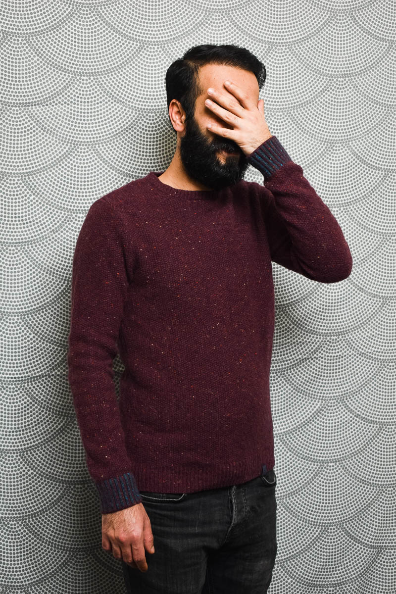 Portrait of refugee Ahmet wearing a maroon sweater and hiding his face with his hand