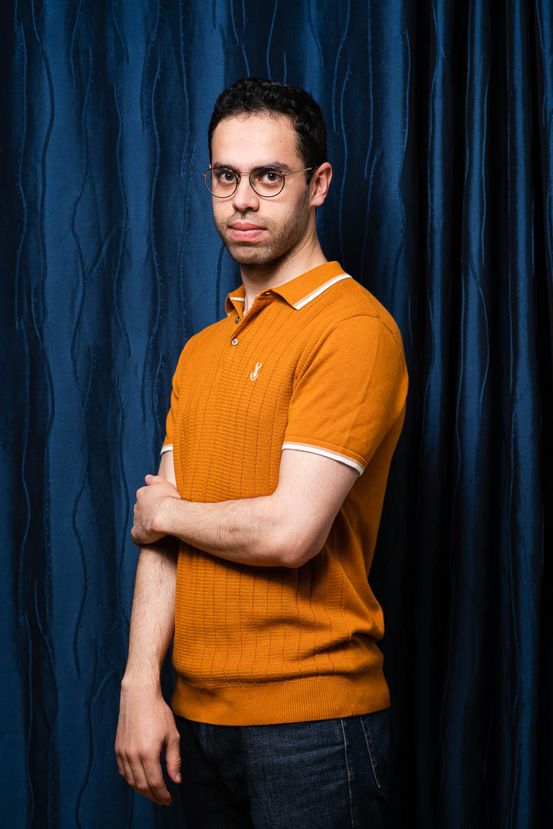 Portrait of refugee Hosam with one hand resting on his other arm standing against a blue curtain