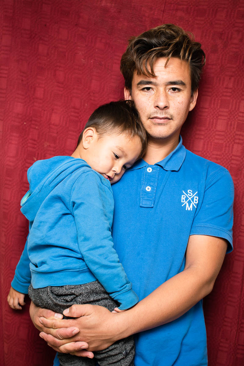 Portrait of refugee Cheraqali wearing a blue polo shirt holding a sleeping toddler in his arms