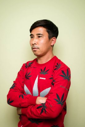 Portrait of refugee Hossinali wearing a red sweater with his arms crossed and looking to the side