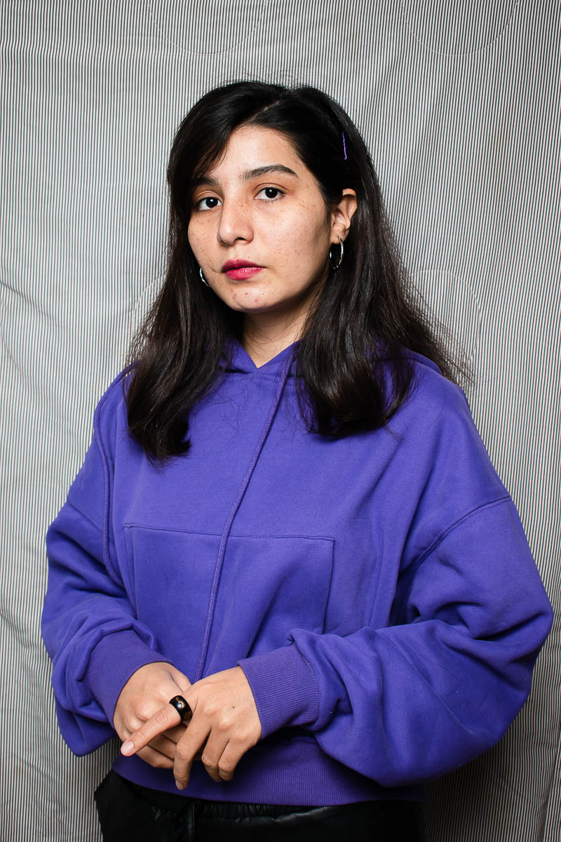 Portrait of refugee Masumeh wearing a blue shirt with her hands rested on her stomach