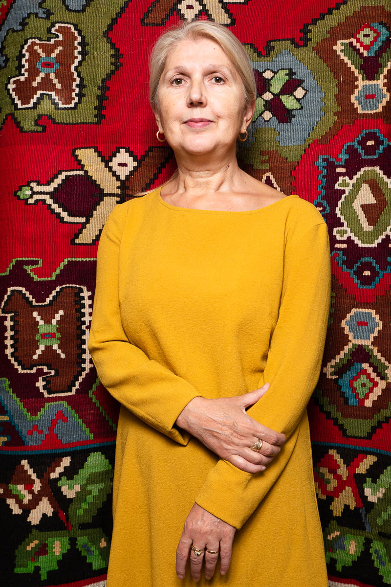 Portrait of refugee Emina with short graying hair wearing a yellow dress standing with her right hand holding her left against an embroidered tapestry