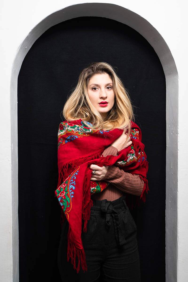Portrait of refugee Inna wrapped around a red Russian shawl standing against a white arch