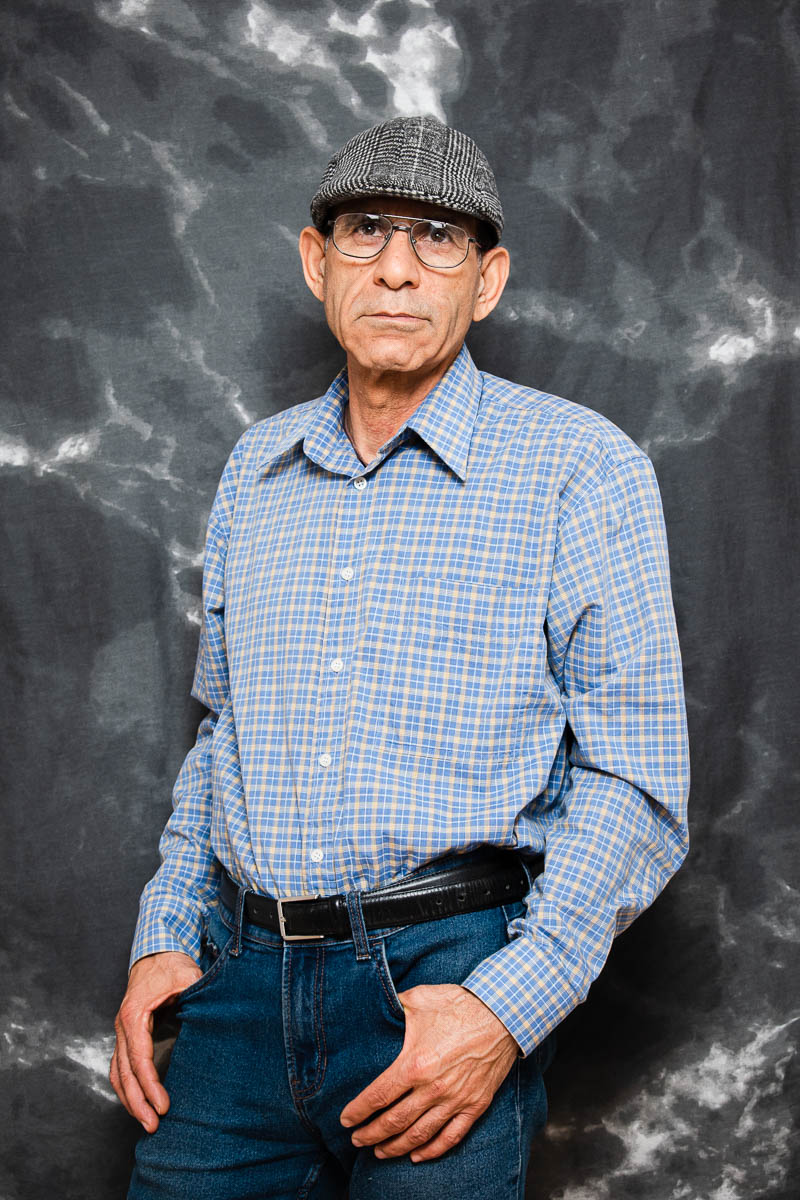 Portrait of refugee Eghbal wearing a plaid shirt and a beret with glasses standing with his thumbs in his jean pockets