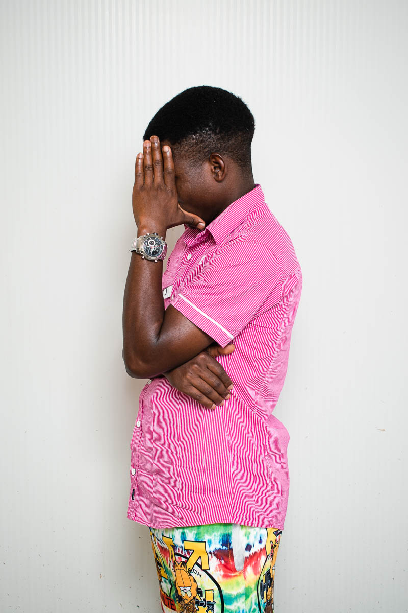 Portrait of refugee Evrard wearing a pink shirt standing sideways and hiding his face with his hand