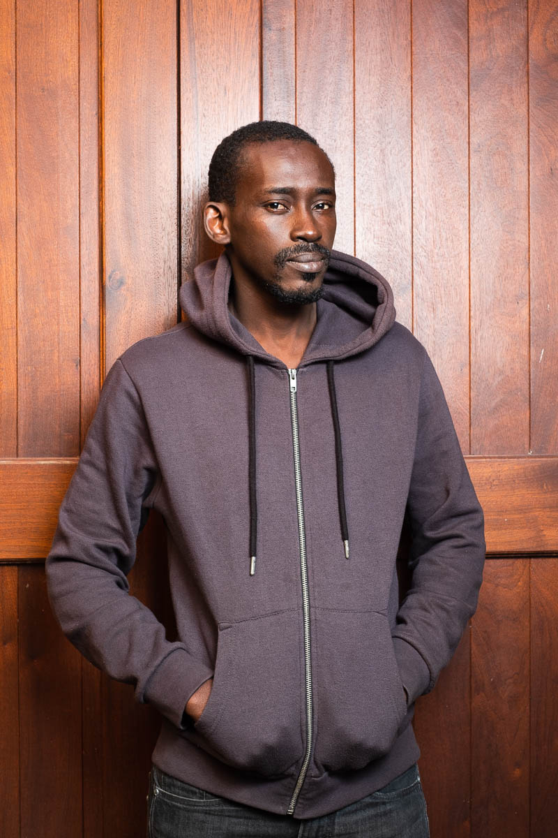 Portrait of refugee Lamine wearing a hoodie with his hands in the pockets standing against a wooden boarded wall
