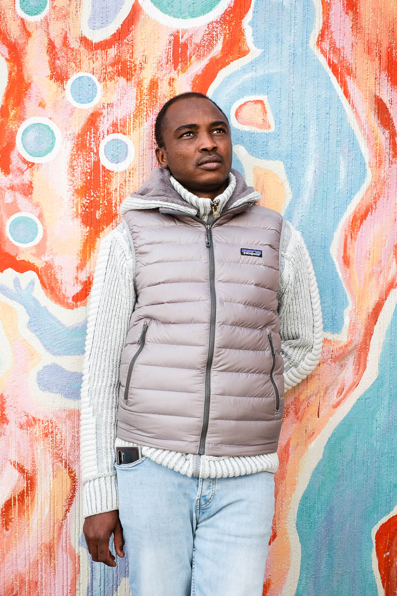 Portrait of refugee Abdallah wearing a puffer jacket standing with one hand behind his back against a painted wall