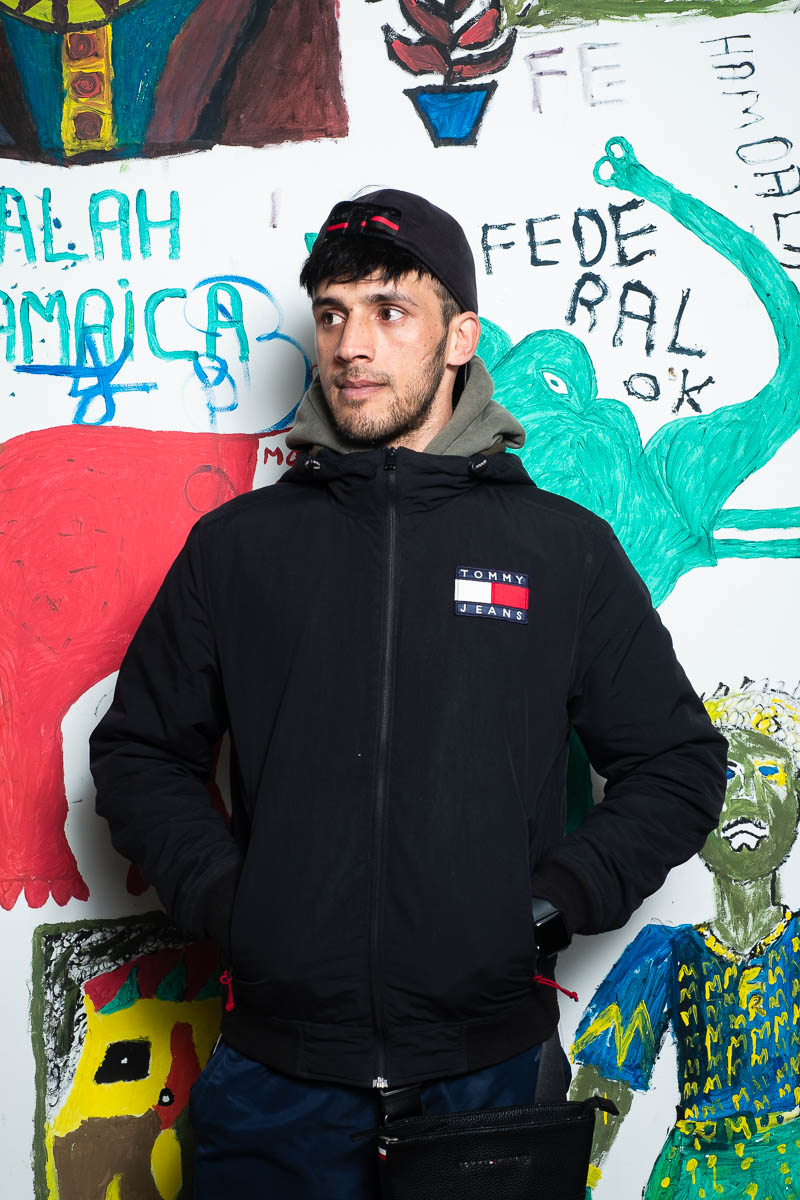 Portrait of refugee Hanib with his hands in the pockets of his zipped hoodie standing against a graffiti wall