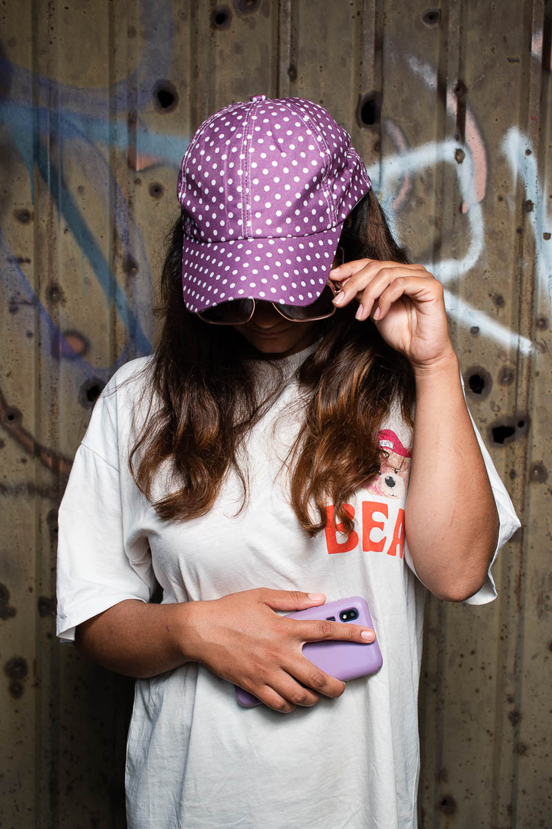 Portrait of refugee Sara hiding her face with a purple polka dot cap and sunglasses while holding a lilac phone in her other hand