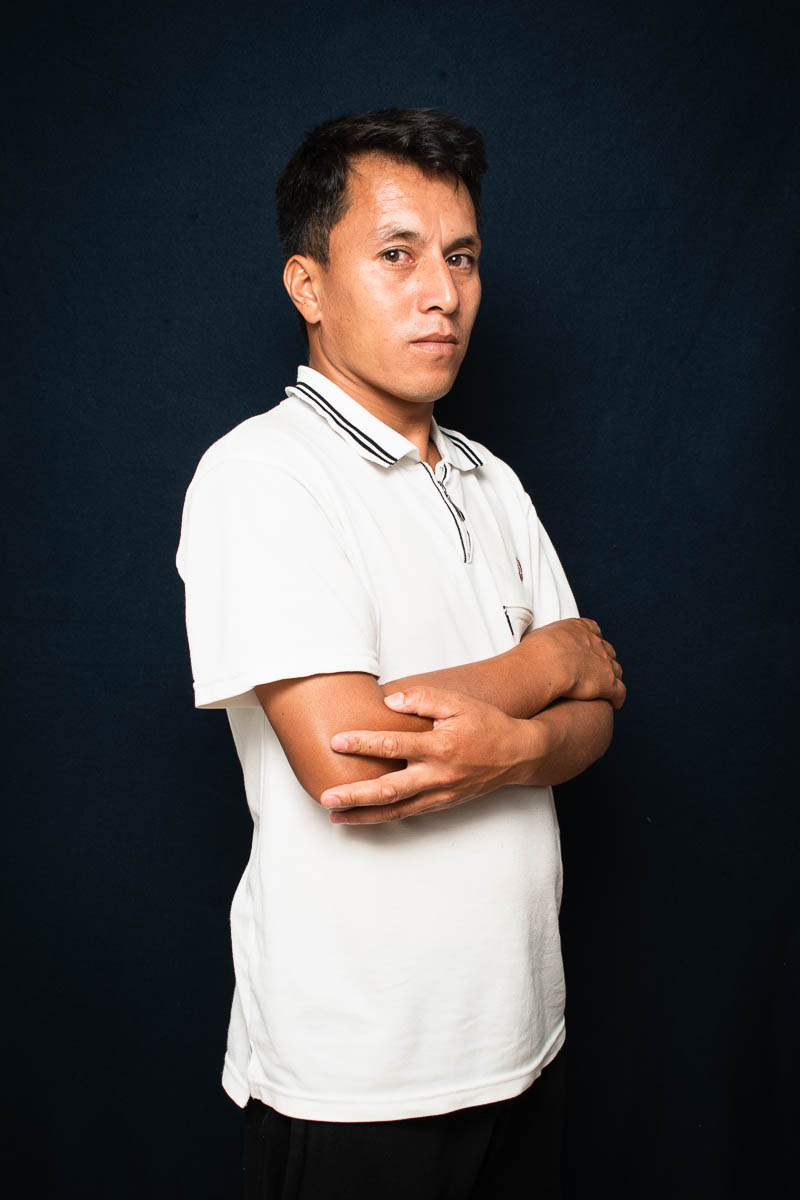 Portrait of refugee Mohamad standing sideways in a white polo shirt with his arms folded