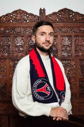 Portrait of refugee Mirza wearing a scarf and stititng with his hands on top of the other on his leg against a wooden partition in the background