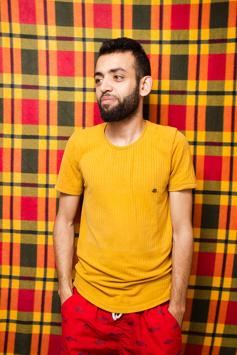 Portrait of refugee Yasir with his hands in the pocket of his red tracks looking to his right and standing against a red and yellow plaid background
