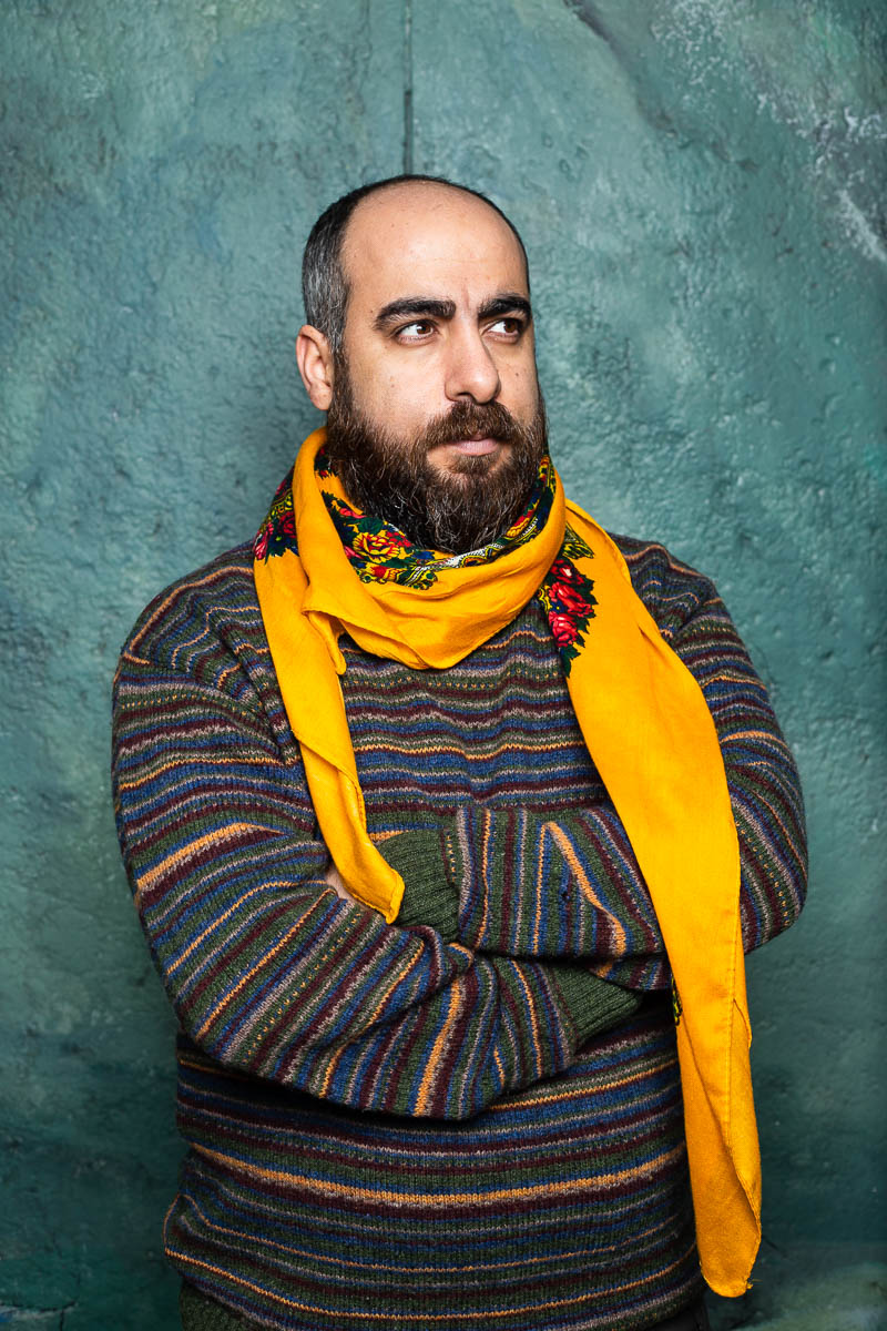 Portrait of refugee Xıdır wearing a sweater and yellow scarf around his neck with his arms crossed looking to his left