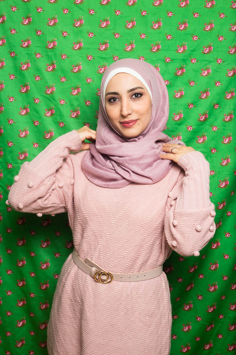 Portrait of refugee Lama wearing a pink dress with both her hands on her shoulder, adjusting her pink hijab and standing against a green background