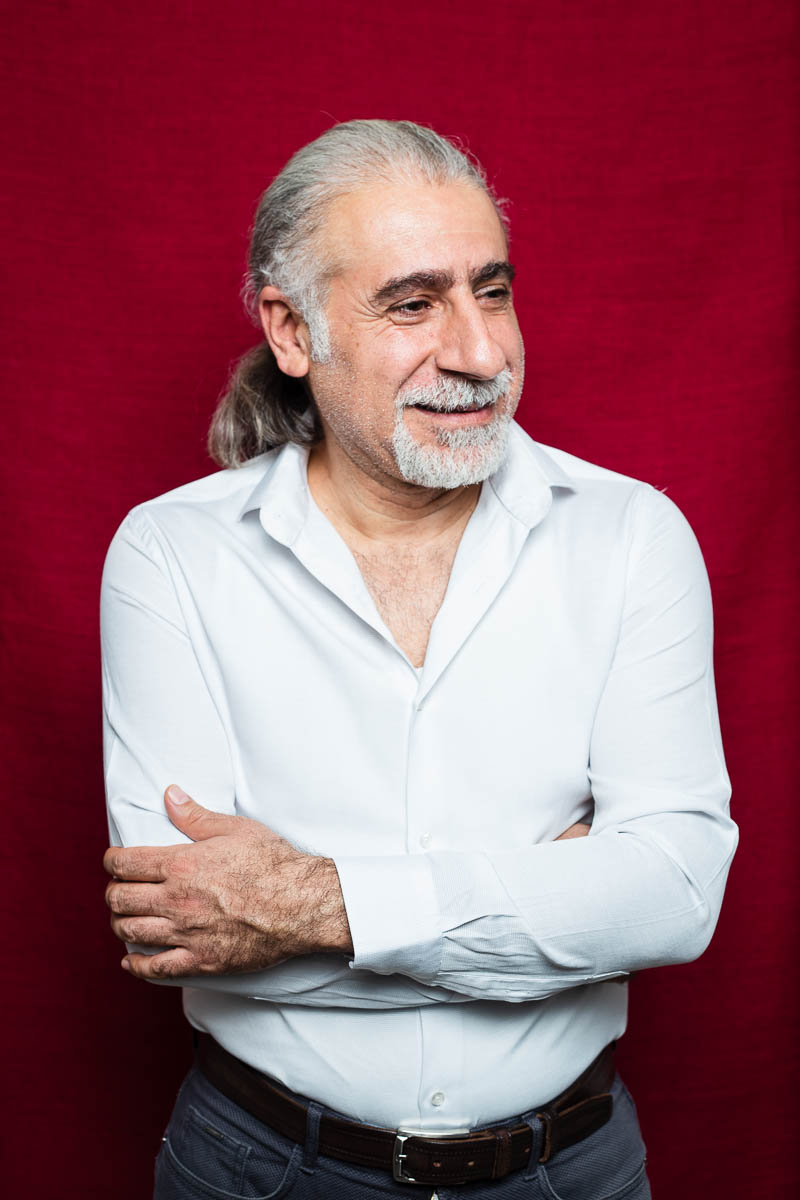 Portrait of refugee Jalal with his hair in a ponytail and hands crossed looking to his left against a red background