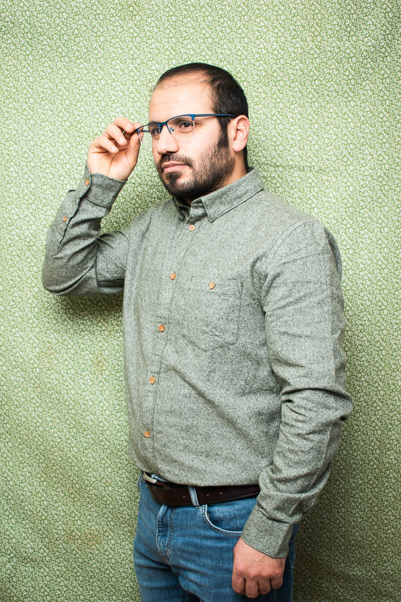 Portrait of refugee Rahmed wearing a green formal shirt with his right hand holding his glasses standing against a green background