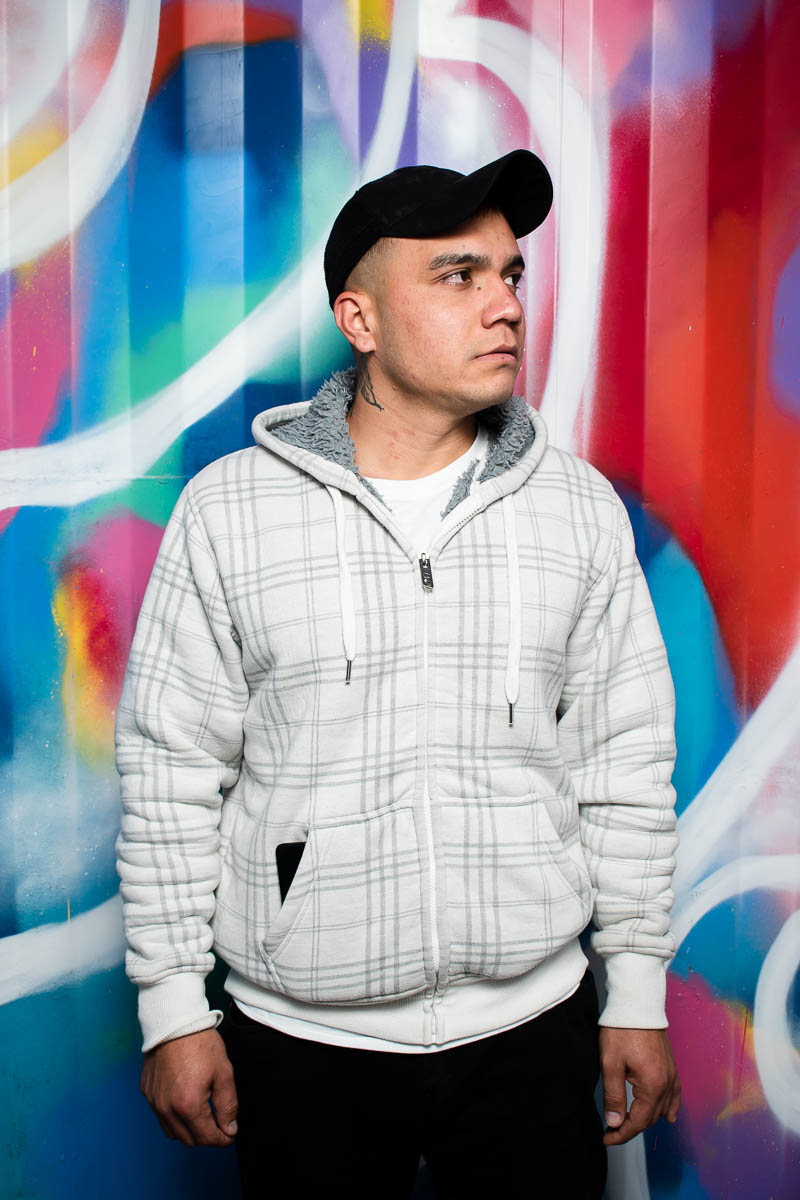 Portrait of refugee Rahman wearing a white hoodie with a black cap looking to his left standing against a graffiti background
