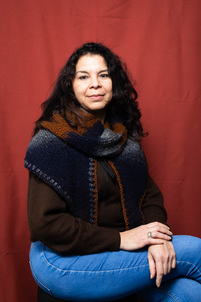 Portrait of refugee Anna with a woolen shawl over her sweater sitting with her hands on top of her folded legs against a red background