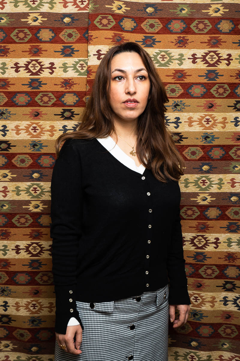 Portrait of refugee Mahtab wearing a black cardigan looking sideways and standing against a woven background