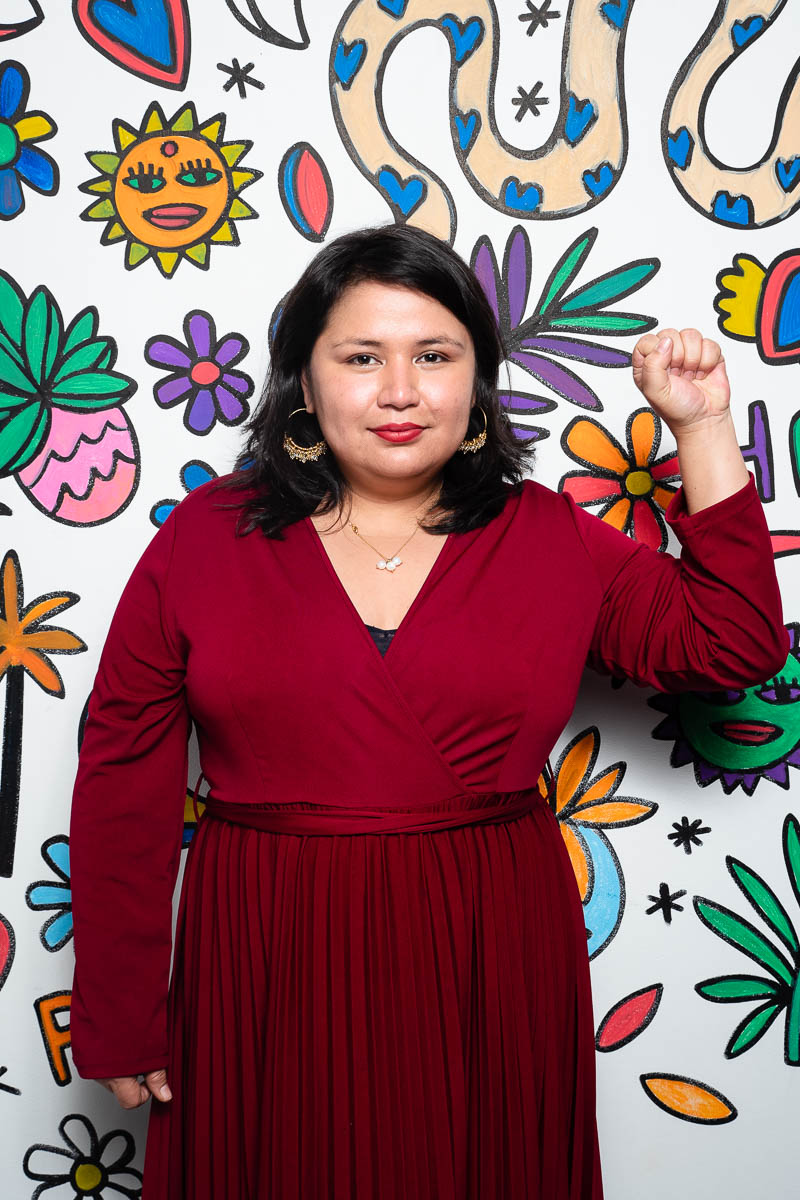 Portrait of refugee Karen wearing a red dress holding her fist up to the left side standing against a wall with colorful doodles