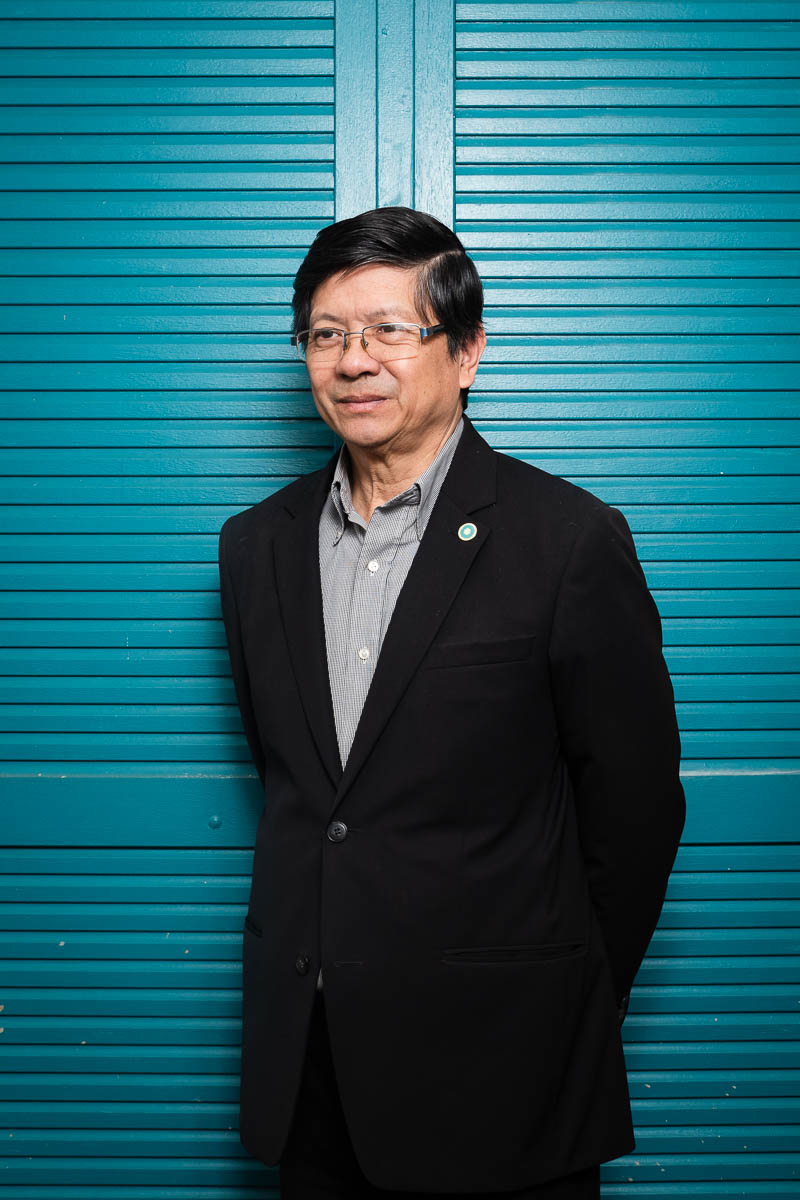 Portrait of refugee Nhan-Dinh wearing a black suit with his hands behind his back standing against a blue paneled door