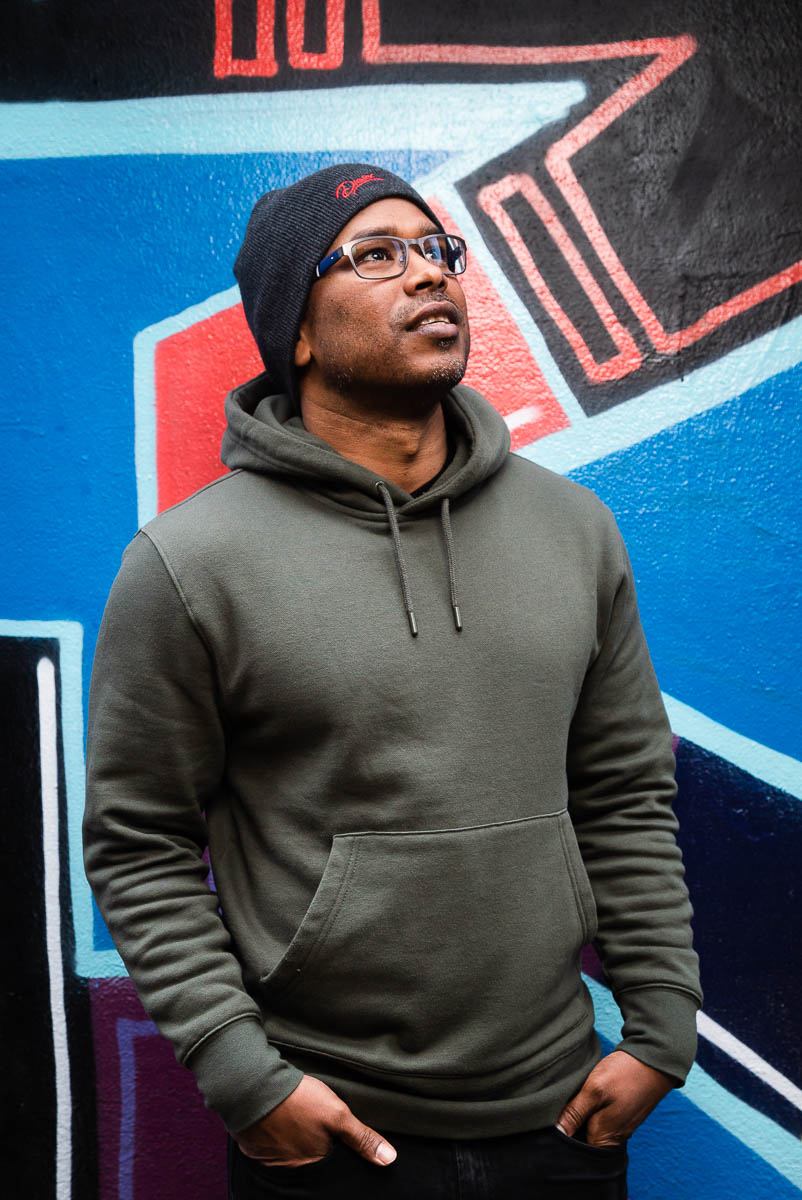 Portrait of refugee Syed wearing a grey hoodie and a beanie with his hands in his pockets standing against a graffiti wall and looking up to the left