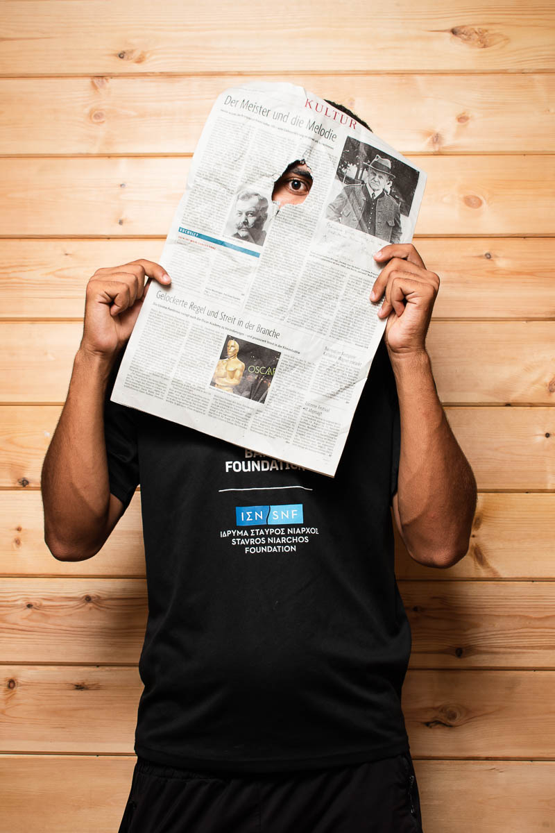 Portrait of refugee Ismail holding a newspaper to cover his face while peeking with one eye through a hole in the newspaper
