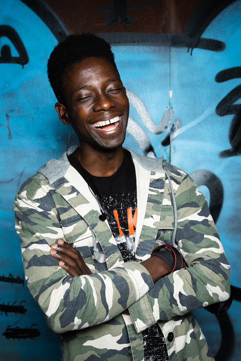 Portrait of refugee Buby wearing a camouflage jacket with his arms crossed and laughing with his eyes closed