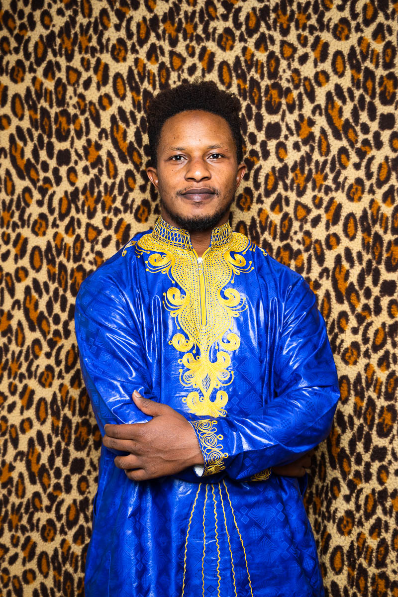 Portrait of refugee Dian wearing a blue dress garb with gold embroidery with his hands crossed standing against an animal print background
