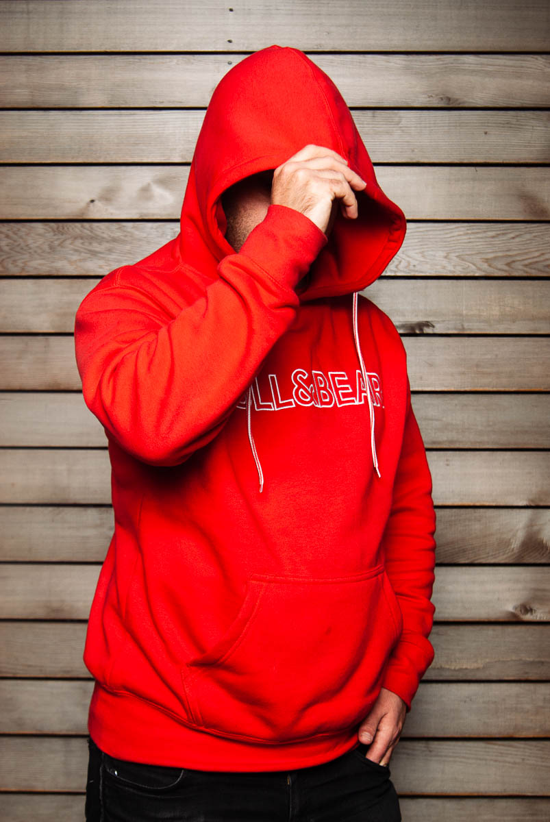 Portrait of refugee Syrus wearing a red hoodie with his left hand in his pocket and his left hand hiding his face while holding the top of the hood