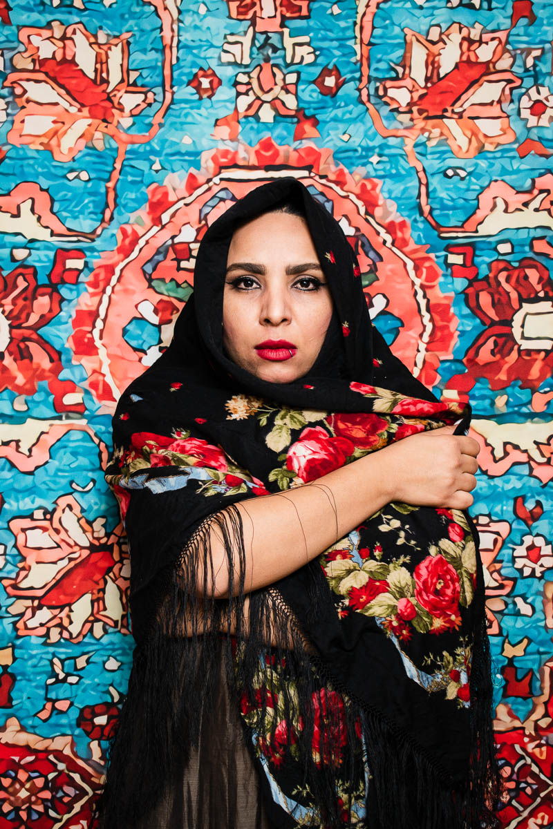 Portrait of refugee Tahmineh wearing a black hijab with a red floral pattern with one hand under the hijan and the other draping it on her shoulder standing against a embroidered tapestry