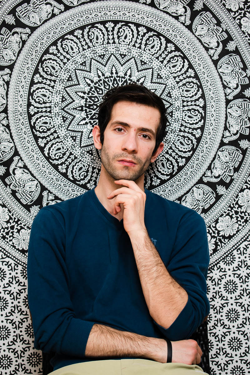 Portrait of refugee 7 with one hand across his waist and the left hand on his chin with a black and white mandala patterned background