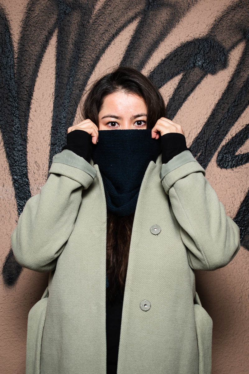Portrait of refugee Setareh wearing a green suede coat and holding a cloth with both her hands to hide the lower half of her face