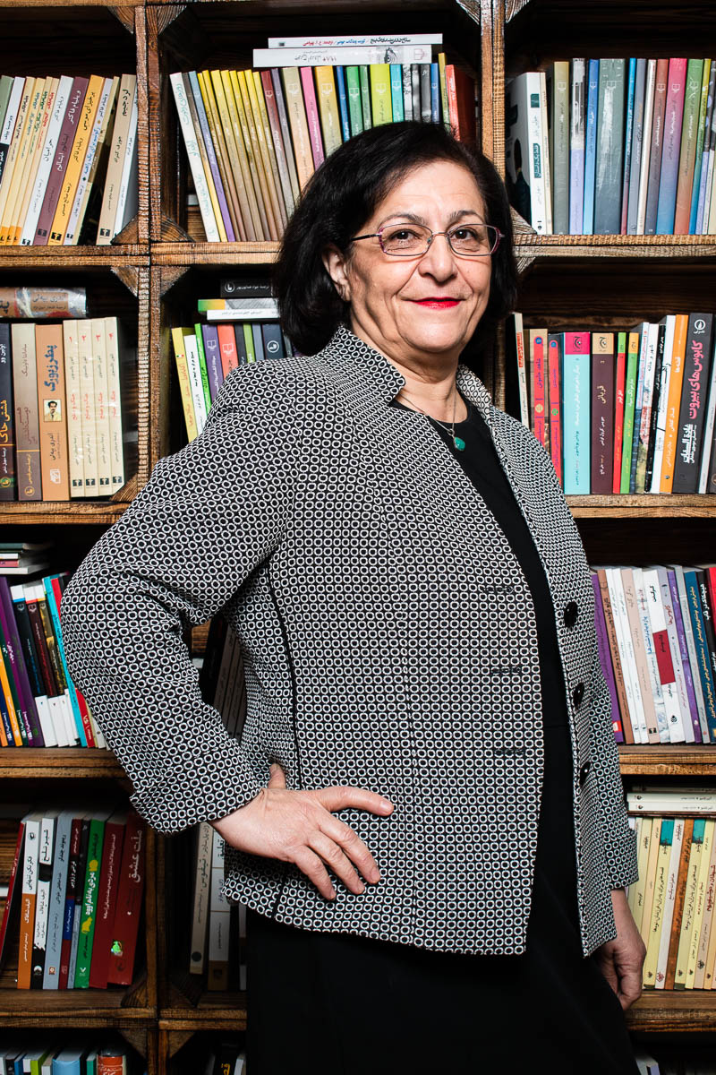 Portrait of refugee Mina wearing a checkered blazer with her hands on her hips standing against a bookshelf