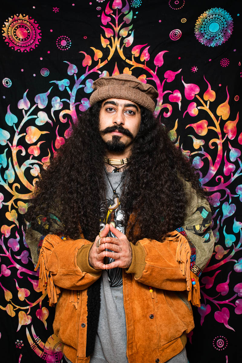 Portrait of refugee Sahand with his long curly hair open wearing a traditional pakol hat standing against a colourful patterned background