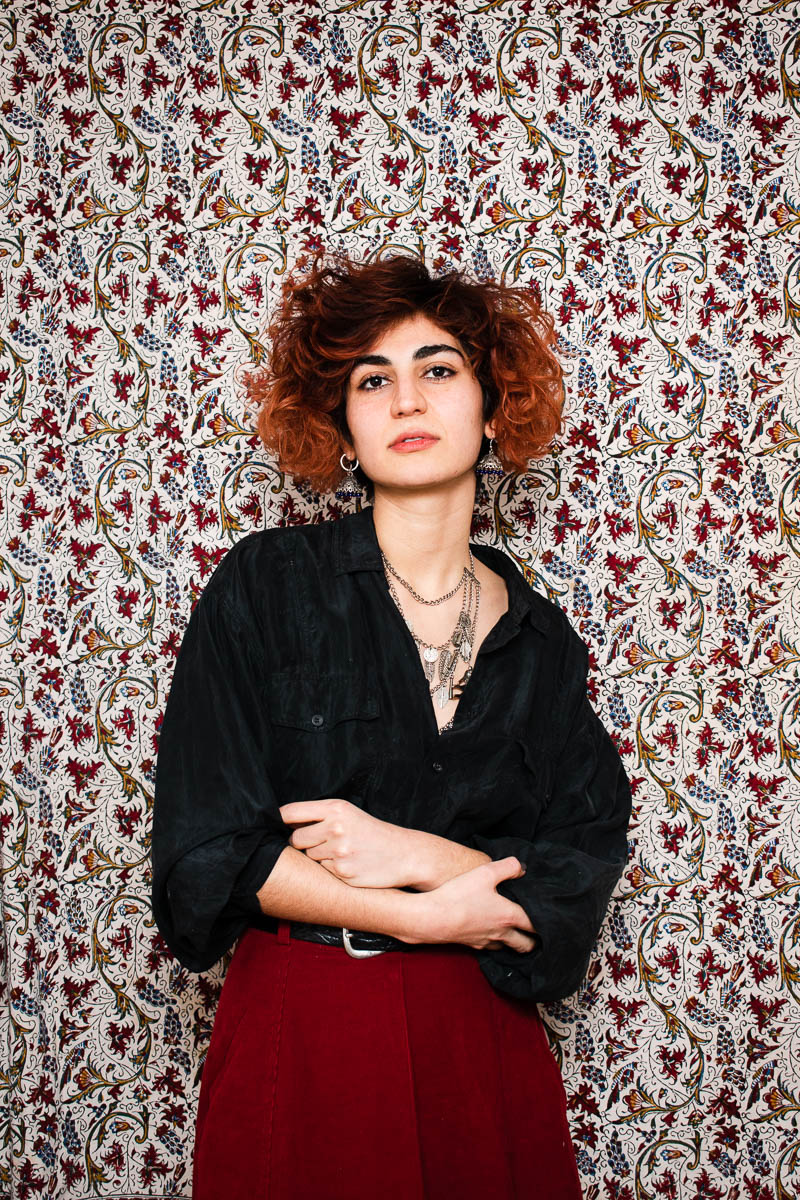 Portrait of refugee Najmeh wearing silver jewelry with short red hair holding the sleeves of her shirt with her hands